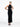 A female model wearing a sleek, black, ribbed yvy dress with a structured leather harness detail, standing in a minimalist white space. She is wearing pointed black heels