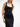 A female model wearing a sleek black square neck dress with double-buckle leather harness detail with straps and gold hardware, standing in a minimalist white space.