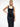 A female model wearing a sleek black square neck dress with studded leather harness detail, standing in a minimalist white space.