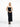 A female model wearing a sleek, black, strapless yvy tube midi dress with a fitted silhouette, standing in a minimalist white space. She is wearing pointed black heels